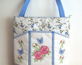 May include: A quilted tote bag featuring a white fabric with blue floral patterns and a front pocket accented with blue gingham trim. The bag displays embroidered pink roses and blue butterflies, with two blue fabric straps and two small blue buttons.