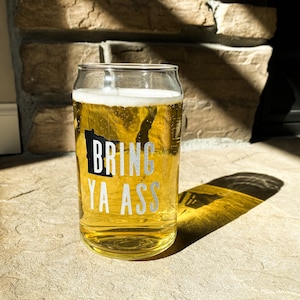 May include: A glass beer can with the text "BRING YA ASS" printed on it. The can is filled with a light yellow beer and is sitting on a stone surface.