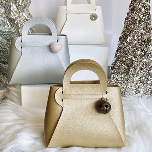 May include: Three decorative handbags in silver, gold, and white, each with a handle and a unique charm. The silver bag has a pink pom-pom, the gold bag has a bell, and the white bag has a deer emblem. The bags are displayed on a white surface.