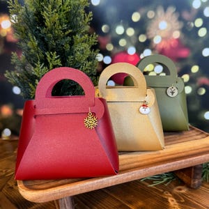May include: Three gift bags in red, gold, and olive green, each with a handle and a decorative charm. The red bag has a snowflake charm, the gold bag has a Santa charm, and the olive green bag has a floral charm. The bags are displayed on a wooden tray.