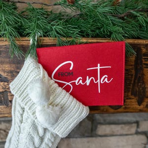 May include: A white knitted stocking with white pom-poms hangs from a wooden mantel. A red felt card with the text "From Santa" is attached to the stocking.