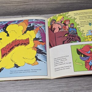 Puede incluir: Ilustraci&oacute;n de un libro abierto, con vi&ntilde;etas de c&oacute;mic. Un panel muestra una explosi&oacute;n amarilla con la palabra "BAWHOOOM". Otros paneles representan a He-Man y sus amigos, y a Orko. El texto incluye di&aacute;logos y narrativa.