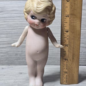 May include: A vintage bisque doll with blonde hair and delicate facial features. The doll has large eyes and is nude. A wooden ruler is next to the doll, measuring approximately 6 inches in height.