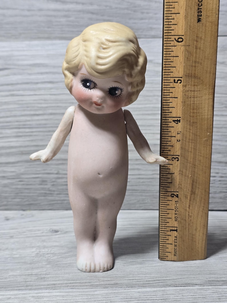 May include: A vintage bisque doll with blonde hair and delicate facial features. The doll has large eyes and is nude. A wooden ruler is next to the doll, measuring approximately 6 inches in height.