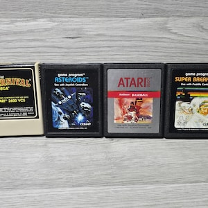 Puede incluir: Cuatro cartuchos de videojuegos Atari 2600 alineados horizontalmente. El primero es un cartucho beige de Carnival, seguido de cartuchos negros para Asteroids, Baseball y Super Breakout. Cada etiqueta presenta títulos y gráficos específicos.