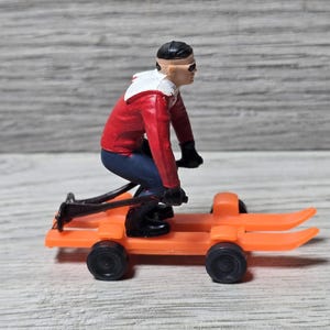 Puede incluir: Un juguete vintage de un esquiador con chaqueta roja, pantalones azules y botas negras, montado en un trineo de esquí naranja con ruedas negras. El esquiador lleva un casco y gafas negros.