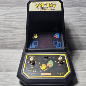 Może przedstawiać: Czarna stołowa gra zręcznościowa Pac-Man firmy Coleco z szyldem z logo Pac-Man, ekranem z grafikami duchów oraz panelem sterowania z dwoma białymi joystickami, przełącznikami wyboru gry oraz żółtymi i pomarańczowymi nadrukami na obudowie.