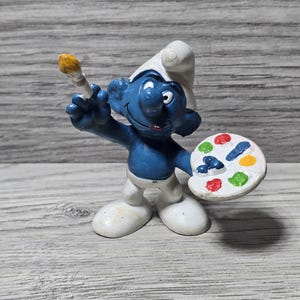 Puede incluir: Figura de un Pitufo azul con pantalón y gorro blancos, sosteniendo un pincel y una paleta de pintura con varios colores. La figura está sobre una superficie de madera.