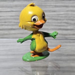 May include: A small, vintage-style figurine of a cartoon duckling with a yellow head and body, green wings, and orange feet and beak. The duckling stands on a green oval base with its arms outstretched and one leg lifted in a playful pose.