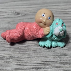 Puede incluir: Una figura vintage de un bebé con un mono rosa abrazando a un perro turquesa. El bebé tiene tez clara, ojos azules y una expresión neutra. El perro tiene detalles blancos y un diseño cosido.