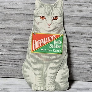 May include: Vintage cat-shaped advertisement for Hoffmann's Reis-Stärke. The cat is gray with orange eyes and a striped pattern. A red and green sign hangs around its neck with the product name and German text.