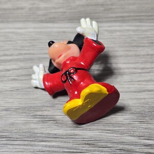 May include: A small figurine of a cartoon mouse in a red outfit with yellow shoes and white gloves. The mouse has a black face and ears, and is posed with its arms raised. The figurine is on a gray wooden surface.