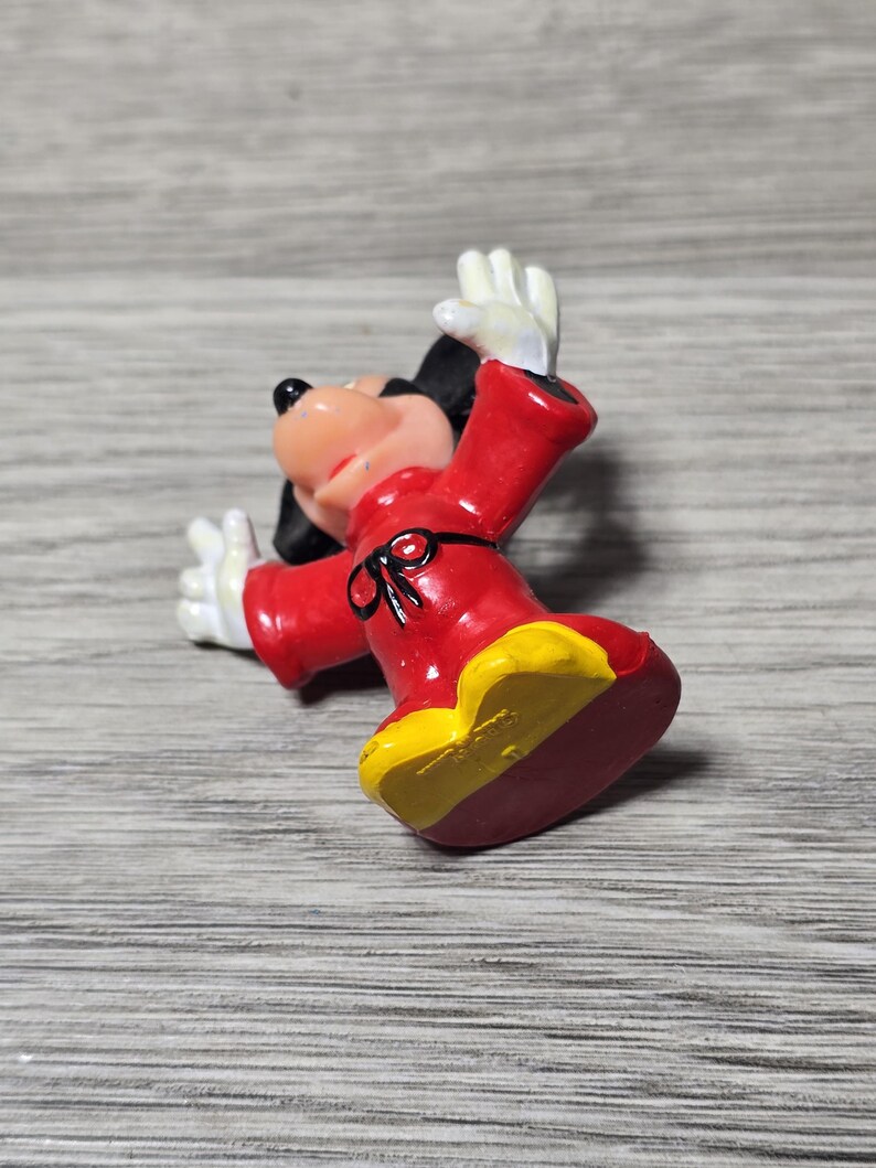 May include: A small figurine of a cartoon mouse in a red outfit with yellow shoes and white gloves. The mouse has a black face and ears, and is posed with its arms raised. The figurine is on a gray wooden surface.