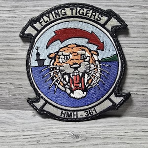 May include: A circular embroidered patch featuring a tiger head in the center with an open mouth, a red arrow above, a silhouette of a ship on the left, and green land on the right. The text FLYING TIGERS is at the top and HMH-361 is at the bottom.