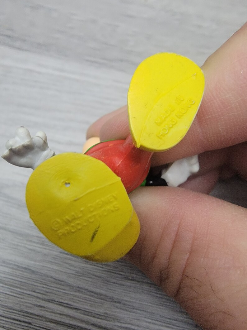 Pode incluir: Figurinha de pl&aacute;stico amarela de um personagem de desenho animado com roupas vermelhas, verdes e brancas. A figurinha tem uma base amarela com o texto "&copy; WALT DISNEY PRODUCTIONS" na parte inferior.