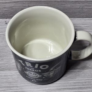 Puede incluir: Una taza de cer&aacute;mica con exterior gris oscuro e interior color crema. La taza presenta el texto "TRIO of TERROR" e ilustraciones de dibujos animados. El asa es blanca. Un art&iacute;culo divertido y novedoso.