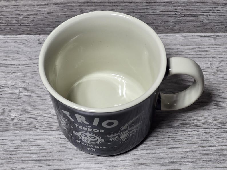 Puede incluir: Una taza de cer&aacute;mica con exterior gris oscuro e interior color crema. La taza presenta el texto "TRIO of TERROR" e ilustraciones de dibujos animados. El asa es blanca. Un art&iacute;culo divertido y novedoso.