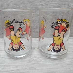 May include: Two clear glass drinking glasses with a Cabbage Patch Kids design. The glasses feature a cartoon image of a child with a yellow shirt and red overalls, holding a jump rope. The text "Cabbage Patch Kids" is printed in a black circle around the child.