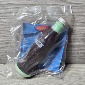 Könnte beinhalten: Ein braunes Spielzeug in Form einer Coca-Cola-Flasche mit grünen und weißen Akzenten, versiegelt in einer transparenten Plastiktüte. Eine blaue Burger King Werbekarte mit dem Coca-Cola-Logo ist enthalten.