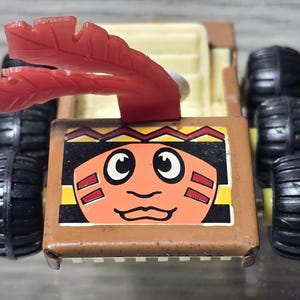 May include: A vintage toy car with a brown body, black tires, and a red feather-like decoration. The front of the car features a cartoon face with orange skin, black eyes, and a red and yellow headdress.