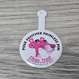 May include: A white, circular Pink Panther Painless Pin with a pink cartoon panther illustration. The pin also features the text "clean 'n treat" and "MEDICATED FIRST AID PADS".