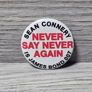 以下が含まれることがあります： 黒文字で「SEAN CONNERY」、赤文字で「NEVER SAY NEVER AGAIN」と書かれた白い丸いボタン。黒文字で「IS JAMES BOND 007」も含まれています。明るい灰色の木目調の背景にボタンが配置されています。
