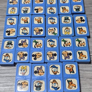 Könnte beinhalten: Eine Sammlung von Vintage-Dominosteinen in Blau mit Disney-Figuren wie Micky Maus, Donald Duck und Goofy. Jeder Dominostein hat einen blauen Rahmen mit einem weißen Zentrum, das eine andere Charakterillustration zeigt. Die Dominosteine sind auf einer hellgrauen Oberfläche angeordnet.
