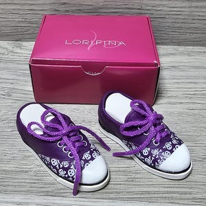 Puede incluir: Un par de zapatillas de deporte moradas con diseños de calaveras y huesos cruzados blancos y cordones morados. Las zapatillas están en una caja rosa con la palabra "LORIFINA" impresa en la parte superior.