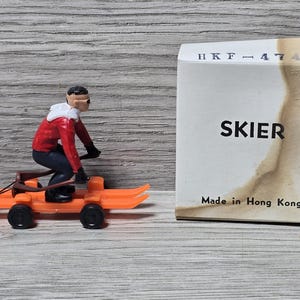 Może przedstawiać: Zabawkowy narciarz w czerwono-białym stroju na pomarańczowych sankach z czarnymi kołami. Narciarz ma na sobie czarny kask i gogle. Na pudełku widnieje napis "SKIER" i "Wyprodukowano w Hongkongu".