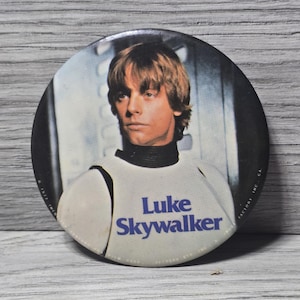 May include: A round button featuring a portrait of Luke Skywalker from Star Wars. The button has a white background with the name "Luke Skywalker" in blue text. The image is set against a gray wooden background.