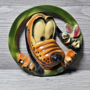May include: Vintage Pluto wall hanging featuring the Disney character in a circular green frame. The ceramic Pluto has a yellow face, orange muzzle, and black nose. A small mouse with red and blue accents is also in the frame.