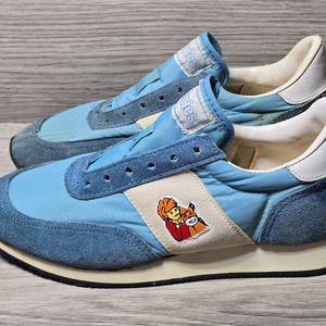 Könnte beinhalten: Paar hellblaue Vintage-Sneaker mit Wildlederakzenten und einem cremefarbenen Streifen mit einer Cartoon-Grafik. Die Sneaker haben eine weiße Sohle und eine blaue Zunge mit dem Aufdruck "Hush".