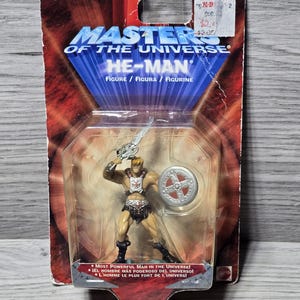 May include: A plastic action figure of He-Man from the Masters of the Universe toy line. He-Man is holding a sword and shield. The figure is packaged in a plastic blister pack with a red, white, and blue background. The packaging includes the text "Most Powerful Man in the Universe!"