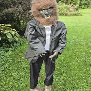 Könnte beinhalten: Eine Werwolf-Figur mit einem Kopf aus braunem Kunstfell, einer detaillierten Maske, gelben Zähnen und spitzen Ohren. Die Figur trägt einen dunkelgrauen Blazer über einem weißen Hemd und einer schwarzen Hose sowie passende fellbedeckte Hände und Füße.