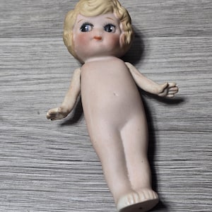 May include: A vintage porcelain doll with a bisque finish. The doll has blonde, curly hair, blue eyes, and rosy cheeks. It is nude, with arms outstretched and feet slightly apart. The doll is lying on a gray wooden surface.