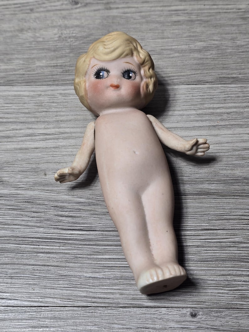 May include: A vintage porcelain doll with a bisque finish. The doll has blonde, curly hair, blue eyes, and rosy cheeks. It is nude, with arms outstretched and feet slightly apart. The doll is lying on a gray wooden surface.
