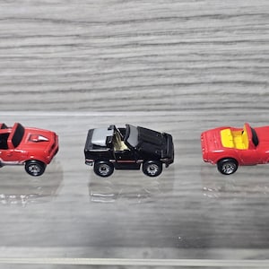 May include: Three miniature toy cars arranged in a row. The left car is red with white side stripes. The middle car is black with a silver roof section. The right car is red with a yellow interior. Each vehicle features four black wheels.