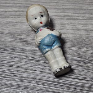Puede incluir: Una peque&ntilde;a mu&ntilde;eca de porcelana vintage con una cara pintada, cabello rubio y pantalones cortos azules. La mu&ntilde;eca tiene un torso y piernas blancos, con una mancha marr&oacute;n en el hombro. La cara de la mu&ntilde;eca tiene labios rojos y ojos negros.