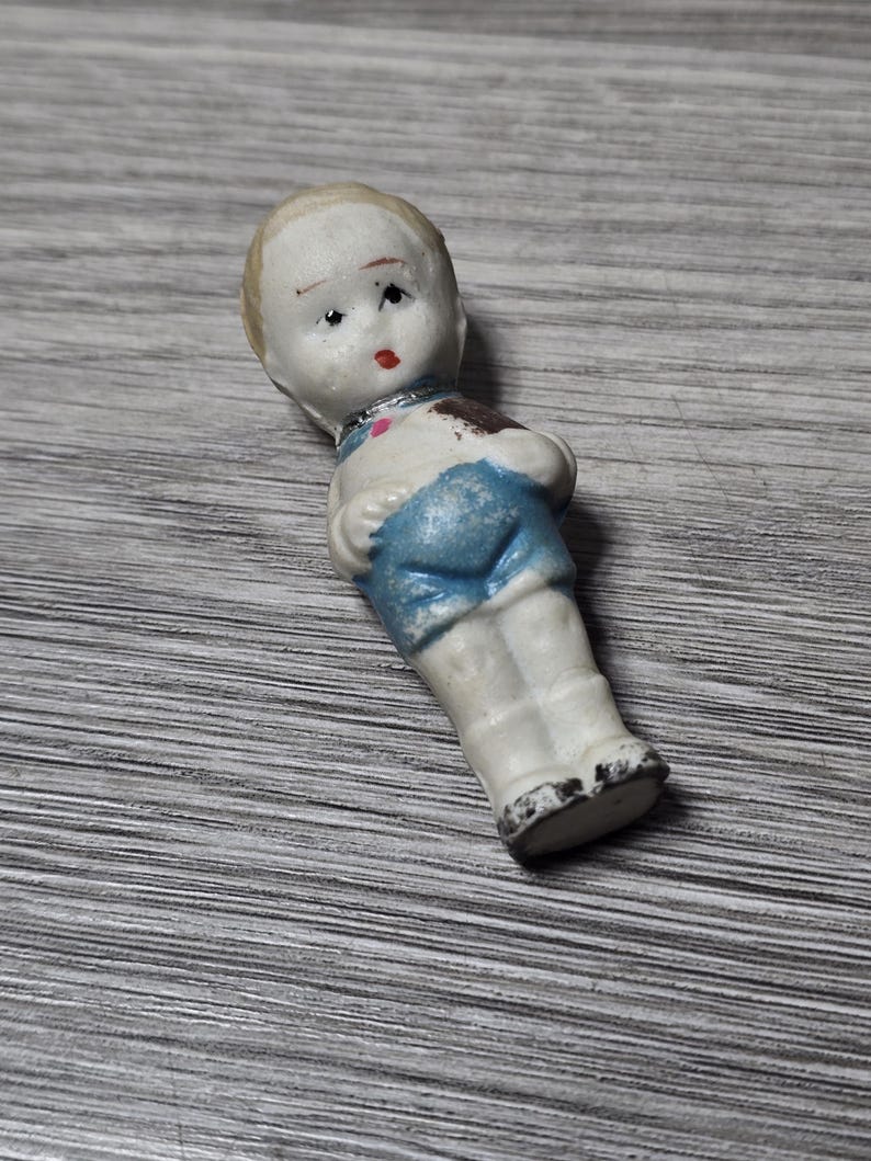 Puede incluir: Una peque&ntilde;a mu&ntilde;eca de porcelana vintage con una cara pintada, cabello rubio y pantalones cortos azules. La mu&ntilde;eca tiene un torso y piernas blancos, con una mancha marr&oacute;n en el hombro. La cara de la mu&ntilde;eca tiene labios rojos y ojos negros.