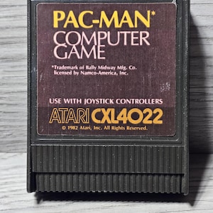 Może przedstawiać: Kasetka do gry komputerowej Atari Pac-Man w stylu vintage. Etykieta zawiera tytuł gry w kolorze żółtym i białym, z logo Atari i informacjami o prawach autorskich. Kasetka jest czarna.