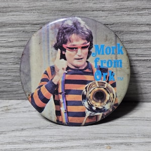 May include: A round button featuring a portrait of a man with dark hair, wearing orange-rimmed glasses and an orange and black striped shirt. The text "Mork from Ork" is in blue. The man is holding a brass instrument.