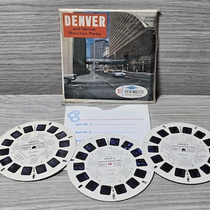 Könnte beinhalten: Vintage View-Master-Set mit dem Titel 'Denver and Denver Mountain Parks'. Die Box zeigt eine Straßenszene. Drei runde Rollen und ein Papiereinleger sind enthalten. Ein nostalgischer Artikel.