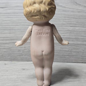 May include: A vintage bisque doll, viewed from the back, with a light skin tone and blonde, curly hair. The doll has outstretched arms and the words "MADE IN JAPAN" are imprinted on its back. The doll is standing on a light gray wooden surface.