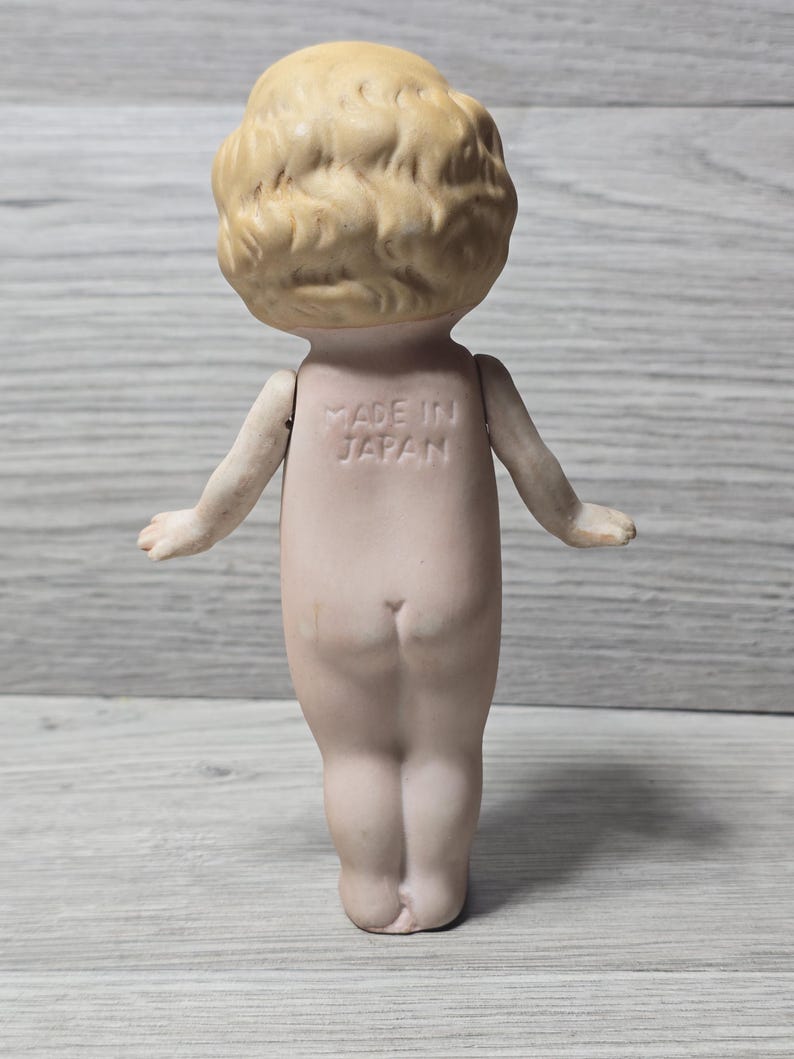 May include: A vintage bisque doll, viewed from the back, with a light skin tone and blonde, curly hair. The doll has outstretched arms and the words "MADE IN JAPAN" are imprinted on its back. The doll is standing on a light gray wooden surface.