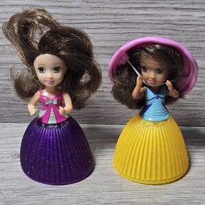 Puede incluir: Dos pequeñas muñecas con cabello castaño y caras pintadas. Una muñeca lleva un top rosa y verde con una falda morada. La otra muñeca lleva un top azul y una falda amarilla, con un sombrero rosa. Las muñecas están sobre una superficie de madera gris.
