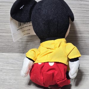 K&ouml;nnte beinhalten: Ein Pl&uuml;sch-Mickey-Mouse-Bohnensackspielzeug, das ein gelbes Hemd und rote Shorts mit wei&szlig;en Punkten tr&auml;gt. Das am Spielzeug befestigte Etikett lautet "Mini Bean Bag Mickey" mit Preisen in kanadischen und US-Dollar.