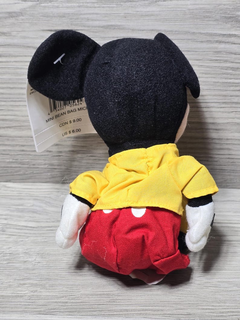 K&ouml;nnte beinhalten: Ein Pl&uuml;sch-Mickey-Mouse-Bohnensackspielzeug, das ein gelbes Hemd und rote Shorts mit wei&szlig;en Punkten tr&auml;gt. Das am Spielzeug befestigte Etikett lautet "Mini Bean Bag Mickey" mit Preisen in kanadischen und US-Dollar.