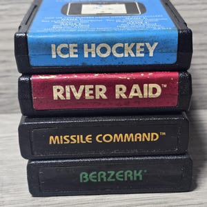 May include: A stack of four black rectangular video game cartridges. The top cartridge has a blue label reading ICE HOCKEY. The second is a red label reading RIVER RAID. The third is a black label with gold text reading MISSILE COMMAND. The bottom is a black label with green text reading BERZERK.