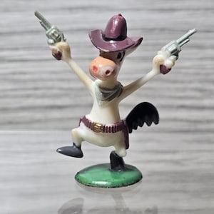 May include: A small figurine of a cartoon donkey wearing a purple cowboy hat, a neckerchief, and a belt with a gold buckle. The donkey is standing on a green circular base, holding a silver-colored toy revolver in each raised hand.