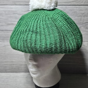 May include: A green knit beret with a white pom-pom on top. The beret is displayed on a white mannequin head against a light gray wood-grain background. The hat is made of a ribbed knit material.